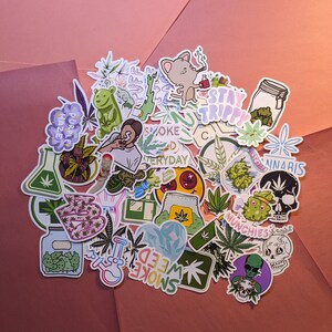 May include: A collection of colorful stickers with cannabis-related designs. The stickers feature illustrations of marijuana leaves, cartoon characters, and text like "Stay Trippy" and "Smoke Weed Everyday". The stickers are arranged on a pink and orange background.
