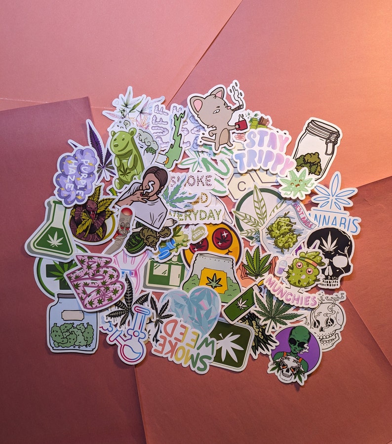 May include: A collection of colorful stickers with cannabis-related designs. The stickers feature illustrations of marijuana leaves, cartoon characters, and text like "Stay Trippy" and "Smoke Weed Everyday". The stickers are arranged on a pink and orange background.
