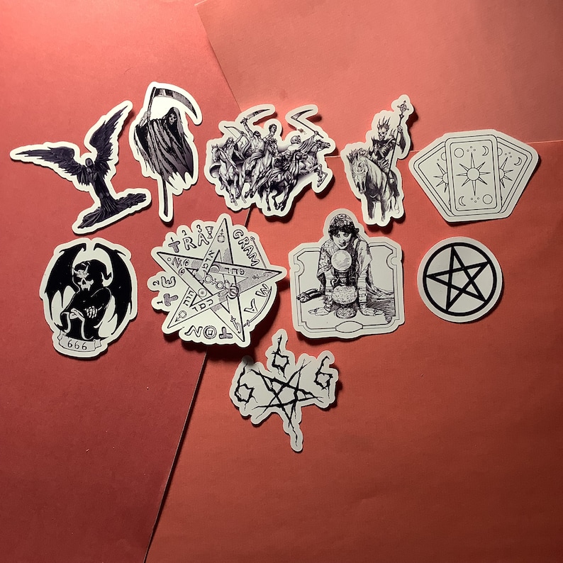 Puede incluir: Una colecci&oacute;n de pegatinas en blanco y negro con temas ocultos y g&oacute;ticos. Los dise&ntilde;os incluyen una parca, una figura alada, un pentagrama, cartas del tarot y otras ilustraciones m&iacute;sticas. Las pegatinas est&aacute;n dispuestas sobre una superficie roja.