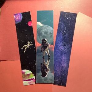 May include: Three rectangular bookmarks with space-themed designs. One shows an astronaut floating in space, another features an astronaut standing before a large moon, and the third depicts a starry night sky with a sun and constellations.