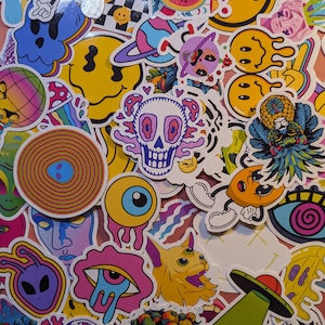 May include: A collection of colorful, psychedelic-themed stickers. Designs include a melting smiley face, a skull with mushroom accents, an eyeball with dripping tears, a cat with a third eye, and a cartoon moon with legs. The stickers are on a pink background.