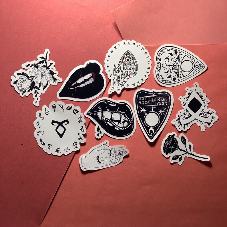 Puede incluir: Una colecci&oacute;n de pegatinas en blanco y negro con temas ocultos y g&oacute;ticos. Los dise&ntilde;os incluyen tableros Ouija, colmillos de vampiro, labios, rosas e ilustraciones de manos. Las pegatinas est&aacute;n dispuestas sobre un fondo rosa.