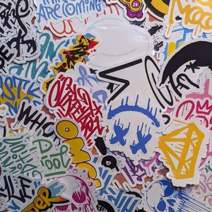 Puede incluir: Una colección de pegatinas coloridas con varios diseños y texto. Las pegatinas presentan arte estilo grafiti, incluyendo palabras, frases y formas abstractas en una variedad de colores como azul, amarillo, rojo y negro. Algunas pegatinas tienen un diseño de calavera.