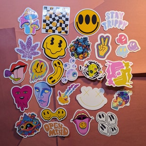 May include: A collection of colorful, psychedelic stickers with various designs, including smiley faces, mushrooms, and trippy graphics. Some stickers feature text like "STAY TRIPPY" and "OPEN MIND". The stickers are arranged on a pink surface.