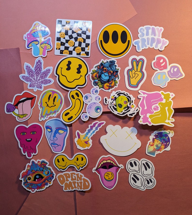 May include: A collection of colorful, psychedelic stickers with various designs, including smiley faces, mushrooms, and trippy graphics. Some stickers feature text like "STAY TRIPPY" and "OPEN MIND". The stickers are arranged on a pink surface.
