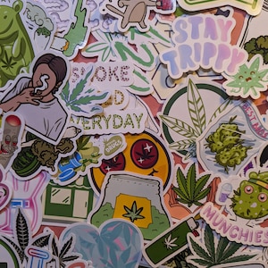 May include: A collection of colorful stickers with cannabis-related designs. The stickers feature images of marijuana leaves, cartoon characters, and phrases like "Stay Trippy" and "Smoke Weed Everyday". The stickers are arranged on a pink surface.