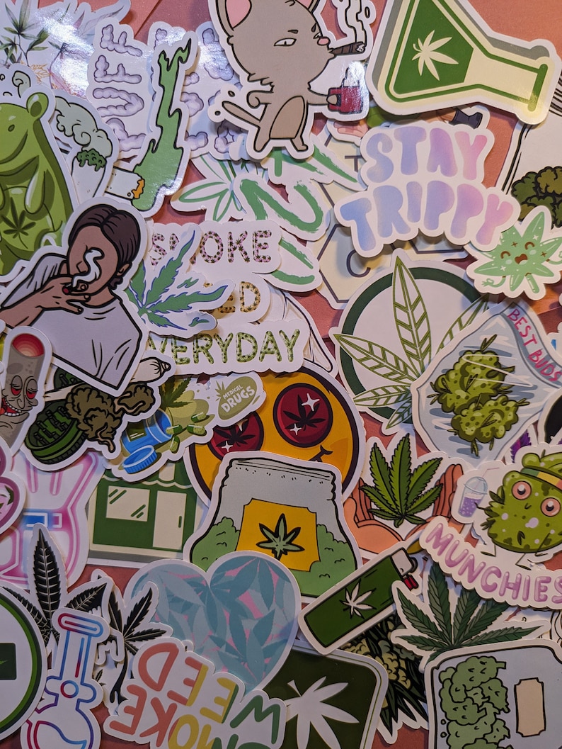May include: A collection of colorful stickers with cannabis-related designs. The stickers feature images of marijuana leaves, cartoon characters, and phrases like "Stay Trippy" and "Smoke Weed Everyday". The stickers are arranged on a pink surface.
