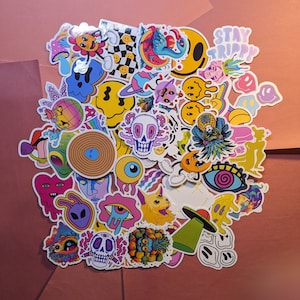 May include: A collection of colorful, psychedelic stickers with various designs, including smiley faces, aliens, skulls, and the text "STAY TRIPPY." The stickers are arranged on a pink surface.