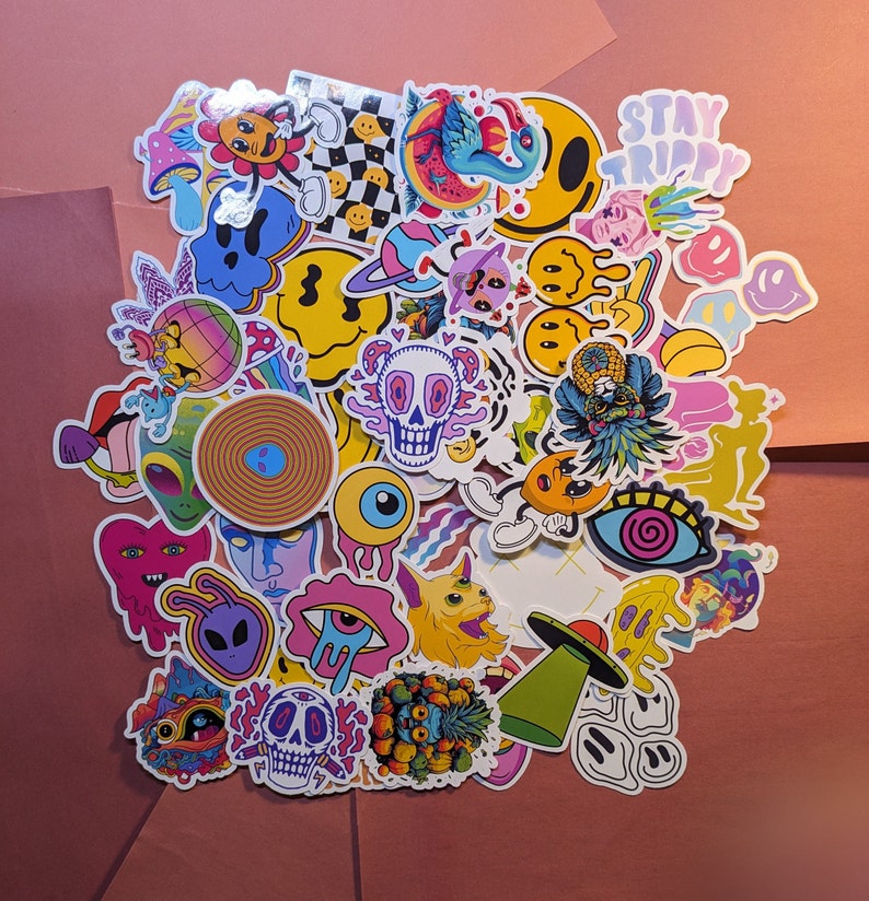 May include: A collection of colorful, psychedelic stickers with various designs, including smiley faces, aliens, skulls, and the text "STAY TRIPPY." The stickers are arranged on a pink surface.
