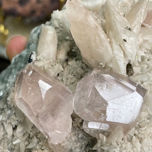 May include: A cluster of white and pink crystals with a rough, natural texture. The crystals are arranged in a variety of shapes and sizes, with some appearing to be translucent. The crystals are set against a dark background.