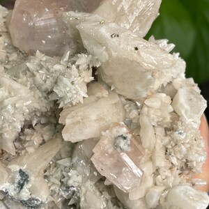 May include: A cluster of white and pink crystals with a rough, natural texture. The crystals are arranged in a variety of shapes and sizes, with some appearing to be translucent. There are small, metallic-looking specks scattered throughout the cluster.