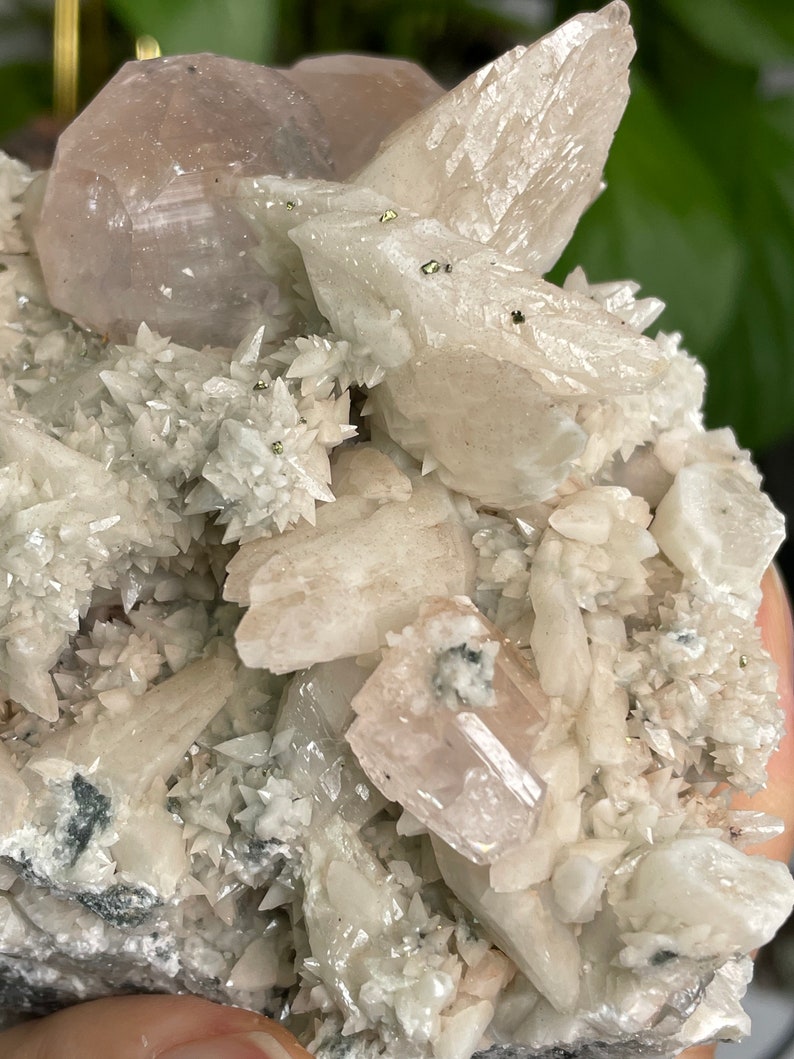 May include: A cluster of white and pink crystals with a rough, natural texture. The crystals are arranged in a variety of shapes and sizes, with some appearing to be translucent. There are small, metallic-looking specks scattered throughout the cluster.