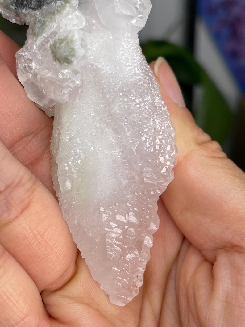 Op de afbeelding: Een cluster van witte kwartskristallen met een groene minerale insluiting. De kristallen zijn doorschijnend en hebben een ruw, getextureerd oppervlak.