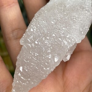 Op de afbeelding: Een witte, doorschijnende kristalkluster met een puntige, grillige vorm. De kristal heeft een ruw, getextureerd oppervlak.