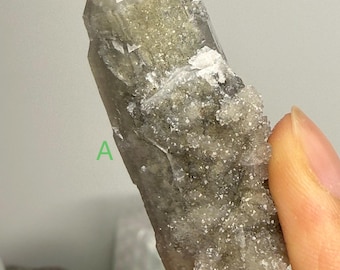 May include: A natural quartz crystal with a pointed tip and a rough, textured surface. The crystal has a translucent, smoky appearance with areas of white and light green. The crystal is held in a hand.