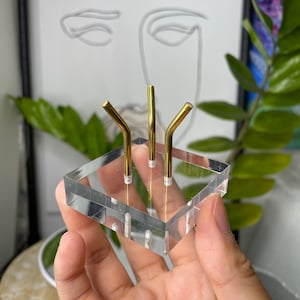 Op de afbeelding: Een transparante acryl ringenhouder met drie gouden metalen pinnen. De houder is ontworpen om meerdere ringen te houden.