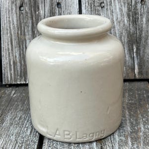 May include: A white ceramic jar with a rounded body and a wide mouth. The jar has a smooth, glazed finish and is embossed with the text "LAB Lagny".