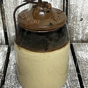 May include: Antique stoneware crock with a dark brown lid and handle. The lid is embossed with the text "WEIR PAT". The crock has a light beige base and a dark brown top. The handle is made of rusty metal.