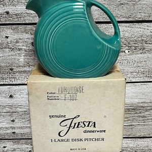 May include: Turquoise Fiesta 1-Large Disk Pitcher. The pitcher is a vibrant turquoise color and has a circular design. The box reads "genuine Fiesta dinnerware" and "Made in USA". The pattern number is F-107.