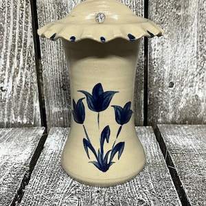 May include: A beige ceramic vase with a flared base and a ruffled lid. The vase is decorated with blue tulip floral designs. A small, clear gem is set in the center of the lid. The vase is on a weathered wooden surface.
