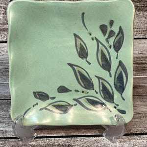 May include: A square ceramic dish with a light green glaze and a black and gray leaf design. The dish is on a wooden surface.