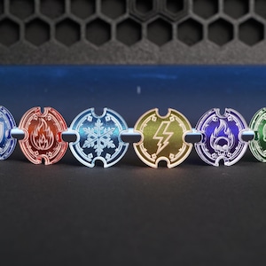 Könnte beinhalten: Ein Set aus sechs runden Acryl-Spielmarken mit eingravierten Elementarsymbolen, darunter ein Schild, eine Flamme, eine Schneeflocke, ein Blitz, ein brennender Totenkopf und ein Totenkopf. Die Marken sind in Blau, Rot, Hellblau, Gelb, Lila und Grün gehalten.