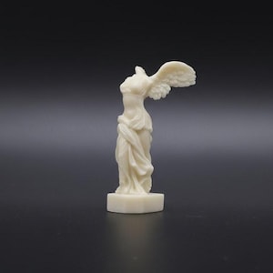 Könnte beinhalten: Eine cremefarbene Statue der Nike von Samothrake, eine hellenistische Skulptur. Die Statue zeigt eine geflügelte Frau mit detaillierten Flügeln und drapierten Kleidern. Die Skulptur ist auf einem kleinen, rechteckigen Sockel montiert.