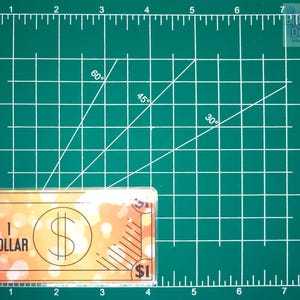 May include: A fake $1 bill with a blurry orange background and the text "1 DOLLAR" printed on it. The bill is on a green cutting mat with white lines and numbers.