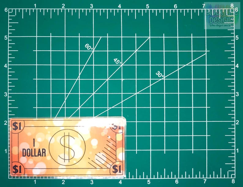 May include: A fake $1 bill with a blurry orange background and the text "1 DOLLAR" printed on it. The bill is on a green cutting mat with white lines and numbers.
