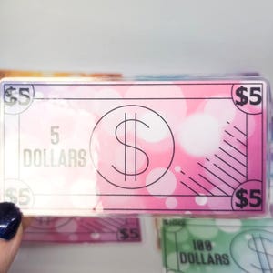 May include: A stack of colorful play money bills with a pink $5 bill on top. The $5 bill has a large dollar sign in a circle and the text "5 DOLLARS" printed on it.