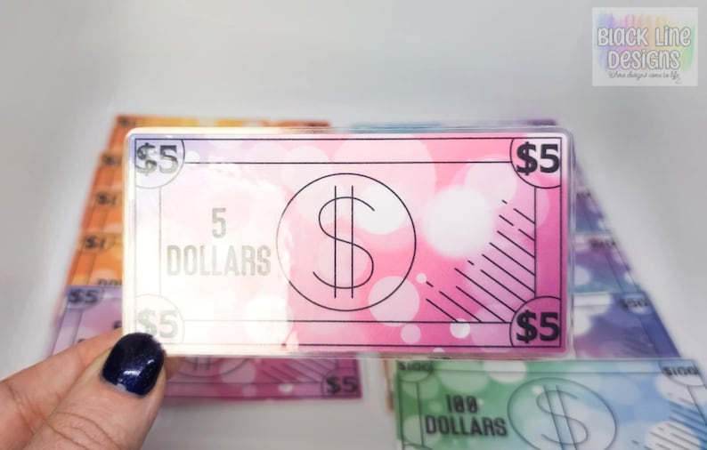 May include: A stack of colorful play money bills with a pink $5 bill on top. The $5 bill has a large dollar sign in a circle and the text "5 DOLLARS" printed on it.