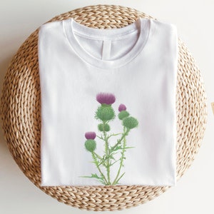 Puede incluir: Camiseta blanca con un estampado gr&aacute;fico de una flor de cardo p&uacute;rpura. La camiseta est&aacute; sobre una estera de paja tejida. Opci&oacute;n de color: Blanco. Boho Tee Sostenible & Vegano.