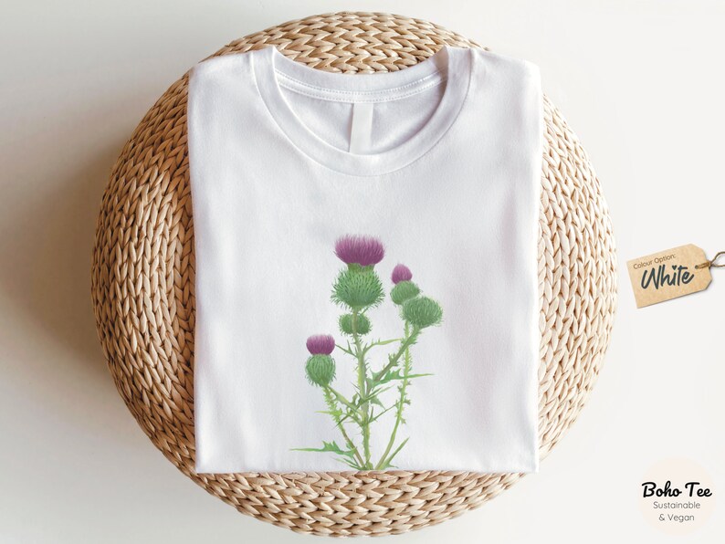 Puede incluir: Camiseta blanca con un estampado gr&aacute;fico de una flor de cardo p&uacute;rpura. La camiseta est&aacute; sobre una estera de paja tejida. Opci&oacute;n de color: Blanco. Boho Tee Sostenible & Vegano.