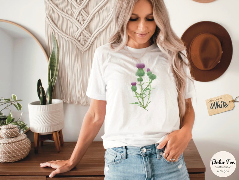 Puede incluir: Camiseta blanca con un gr&aacute;fico de una flor de cardo p&uacute;rpura. La camiseta est&aacute; hecha de un material sostenible y vegano.  "Boho Tee Sustainable & Vegan" est&aacute; impreso en una etiqueta marr&oacute;n.