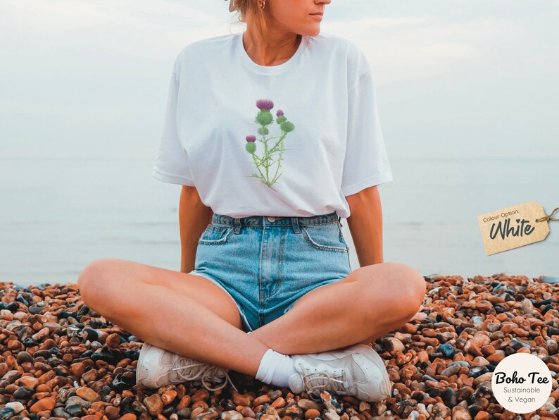 Puede incluir: Una camiseta blanca con un estampado de cardo morado. La camiseta la lleva puesta una persona sentada en una playa con el oc&eacute;ano y el cielo de fondo. El texto "Opci&oacute;n de color: Blanco" est&aacute; en una etiqueta marr&oacute;n en la esquina superior derecha de la imagen. El texto "Boho-Tee Sostenible & Vegano" est&aacute; en un c&iacute;rculo marr&oacute;n en la esquina inferior derecha de la imagen.