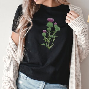 Puede incluir: Una camiseta negra con un gr&aacute;fico de una flor de cardo p&uacute;rpura. La camiseta est&aacute; hecha de materiales sostenibles y veganos. La opci&oacute;n de color es negro.