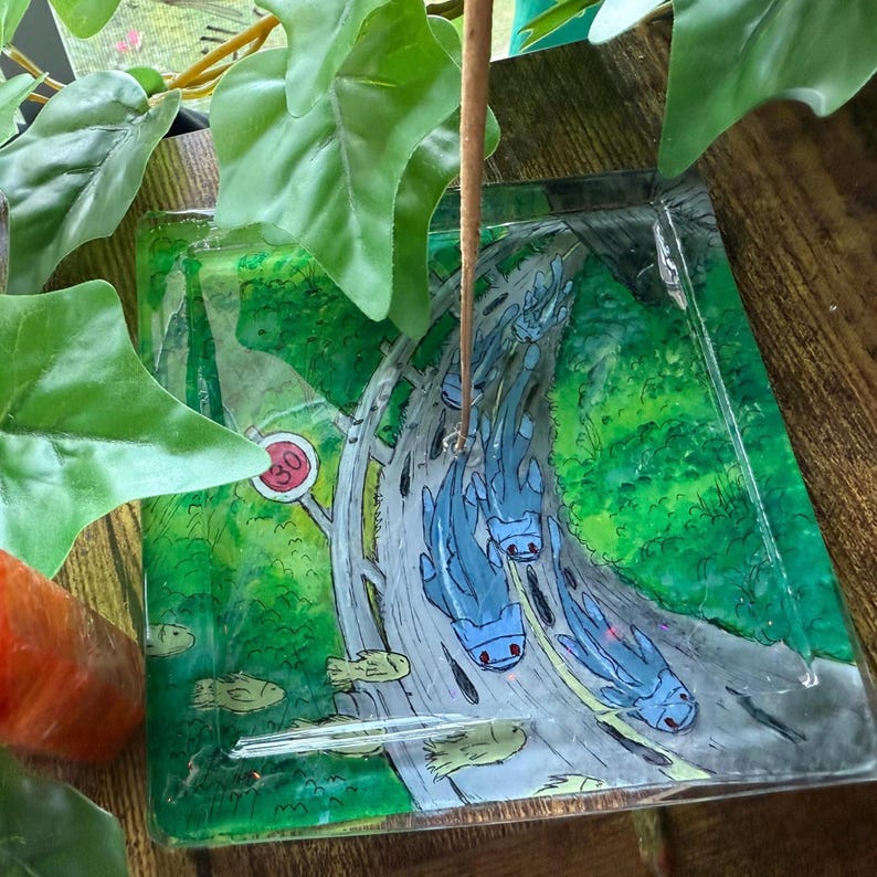 May include: A clear, square glass tray with a road design featuring blue creatures. The road winds through a green forest, with a speed limit sign visible. The tray has a decorative, artistic style.