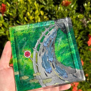 May include: Square resin tray with a hand-painted design featuring a road with blue creatures and a speed limit sign. The tray has a green and gray color scheme, with a glossy finish. The background is blurred with green foliage and red flowers.