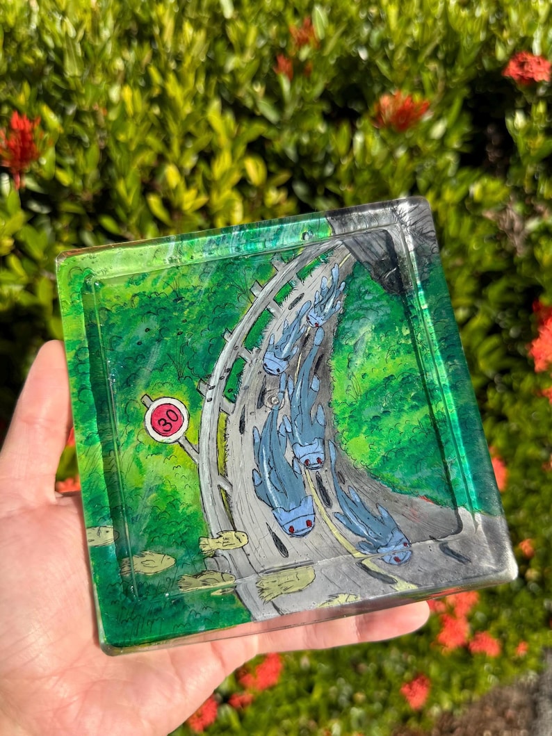 May include: Square resin tray with a hand-painted design featuring a road with blue creatures and a speed limit sign. The tray has a green and gray color scheme, with a glossy finish. The background is blurred with green foliage and red flowers.