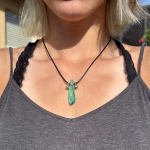 May include: A necklace with a black cord and a green, rectangular pendant. The pendant has a textured appearance and is accented with two small silver beads. The necklace is worn against a gray tank top.