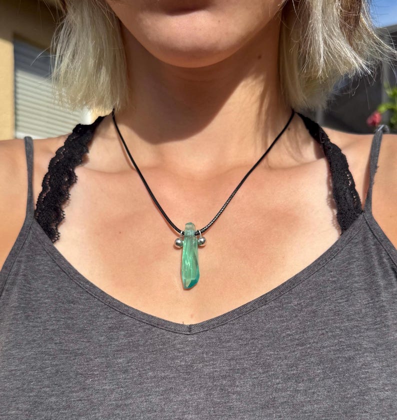 May include: A necklace with a black cord and a green, rectangular pendant. The pendant has a textured appearance and is accented with two small silver beads. The necklace is worn against a gray tank top.