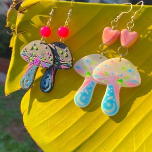 May include: A pair of mushroom-shaped earrings with colorful accents. One pair features gray caps with pink, blue, and green details, while the other has white caps with similar colors. Both pairs have silver hooks and are set against a yellow leaf.