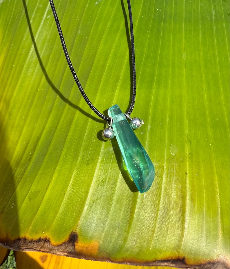 May include: A pendant necklace featuring a translucent, emerald-green crystal pendant suspended from a black cord. The crystal has a faceted shape and is accented with two small, silver-colored beads. The necklace rests on a large, green leaf.