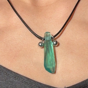 May include: A green crystal pendant necklace. The pendant is a translucent, elongated crystal with a faceted surface. It hangs from a black cord and is accented with two small silver-colored beads. The necklace is worn.
