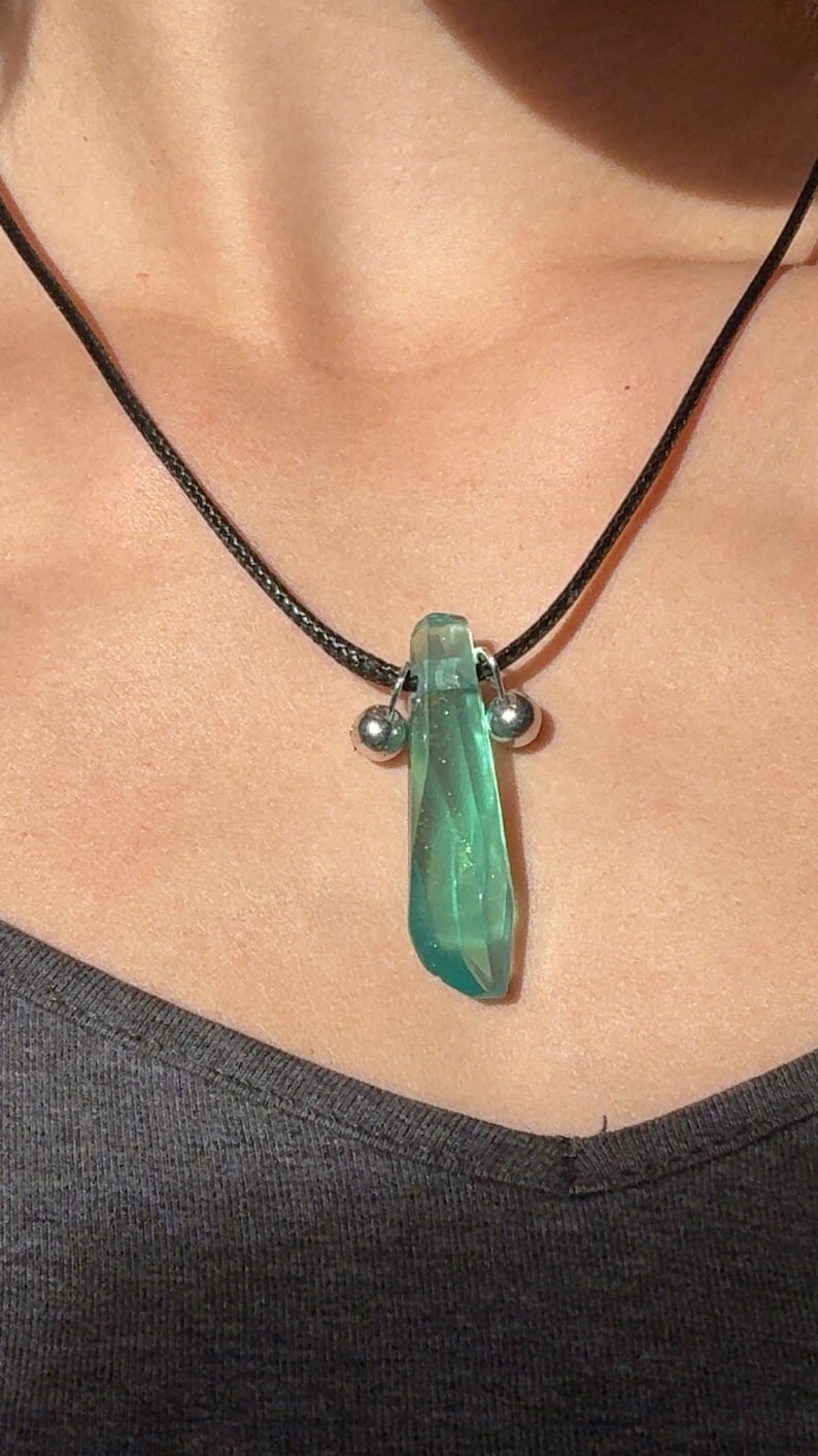 May include: A green crystal pendant necklace. The pendant is a translucent, elongated crystal with a faceted surface. It hangs from a black cord and is accented with two small silver-colored beads. The necklace is worn.
