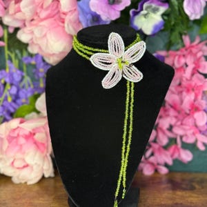 May include: A beaded necklace with a large flower pendant. The flower is pink and white, with green accents. The necklace is displayed on a black mannequin bust, with a backdrop of colorful flowers.
