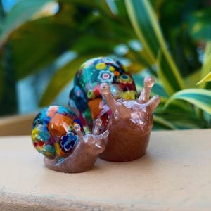 May include: Three glass snail ornaments. Two snails feature colourful, patterned shells with blue, orange, and yellow hues. The third snail is a solid, translucent brown colour. The ornaments are displayed on a light brown surface.