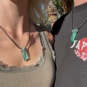 May include: Two people wearing matching necklaces. Each necklace has a light blue, rectangular pendant suspended from a black cord. The pendants are made of a translucent material. The necklaces are worn against a neutral background.