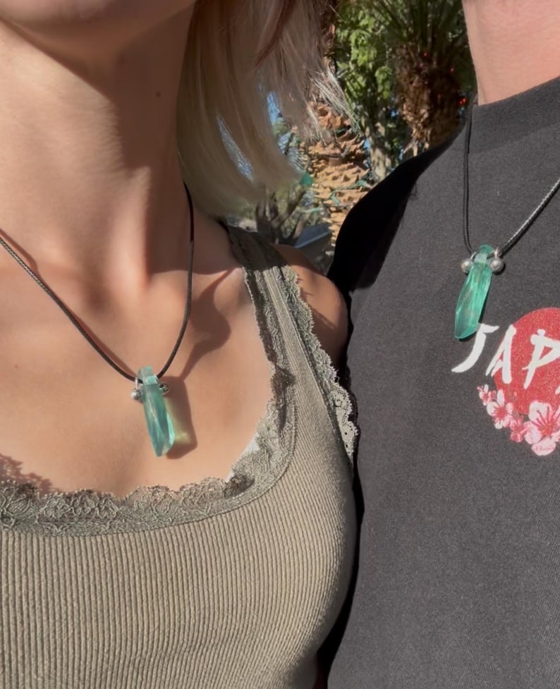 May include: Two people wearing matching necklaces. Each necklace has a light blue, rectangular pendant suspended from a black cord. The pendants are made of a translucent material. The necklaces are worn against a neutral background.