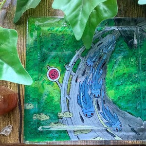May include: Square, transparent coaster with a whimsical design. The coaster features a road scene with blue creatures and a speed limit sign of 30. The background is a vibrant green, with a wooden surface.